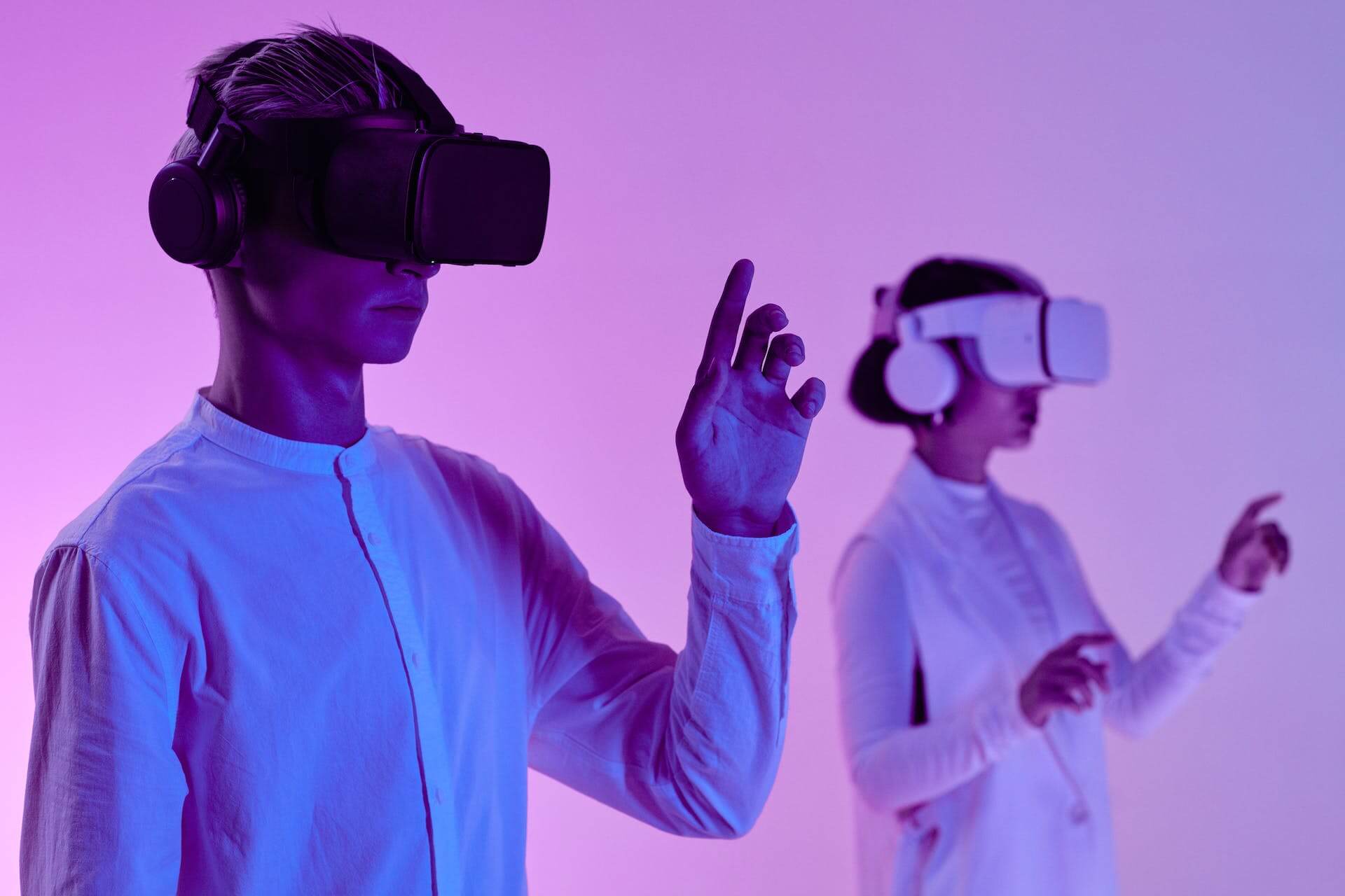 A man and woman with virtual reality goggles. The man is holding up one hand in front of his eyeline. The woman is in the background, slightly out of focus, using both hands to presumably manipulate an interface.