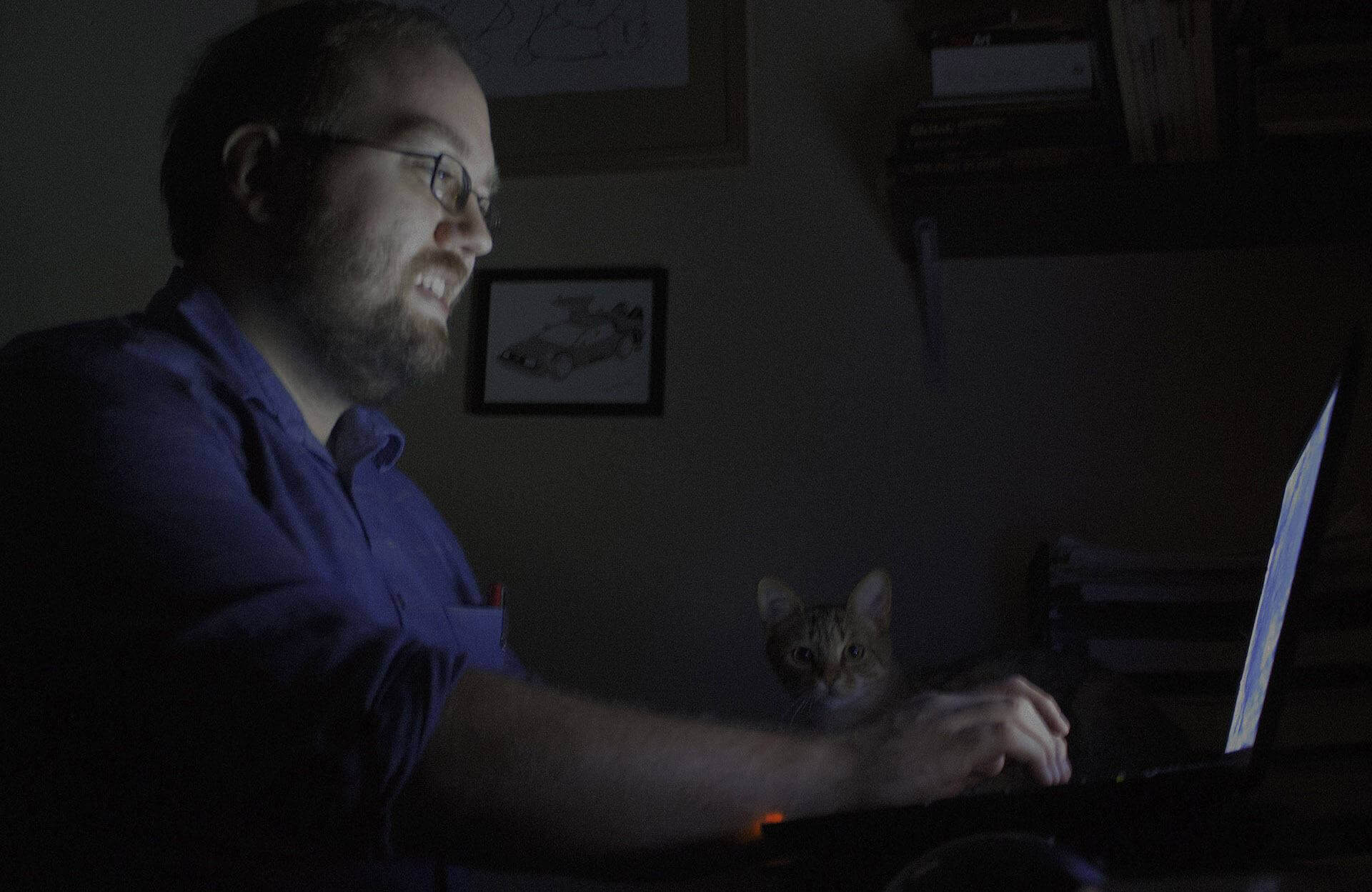 Bearded man with glasses and button-up shirt looking at laptop screen while a kitten is behind him, looking into the camera.